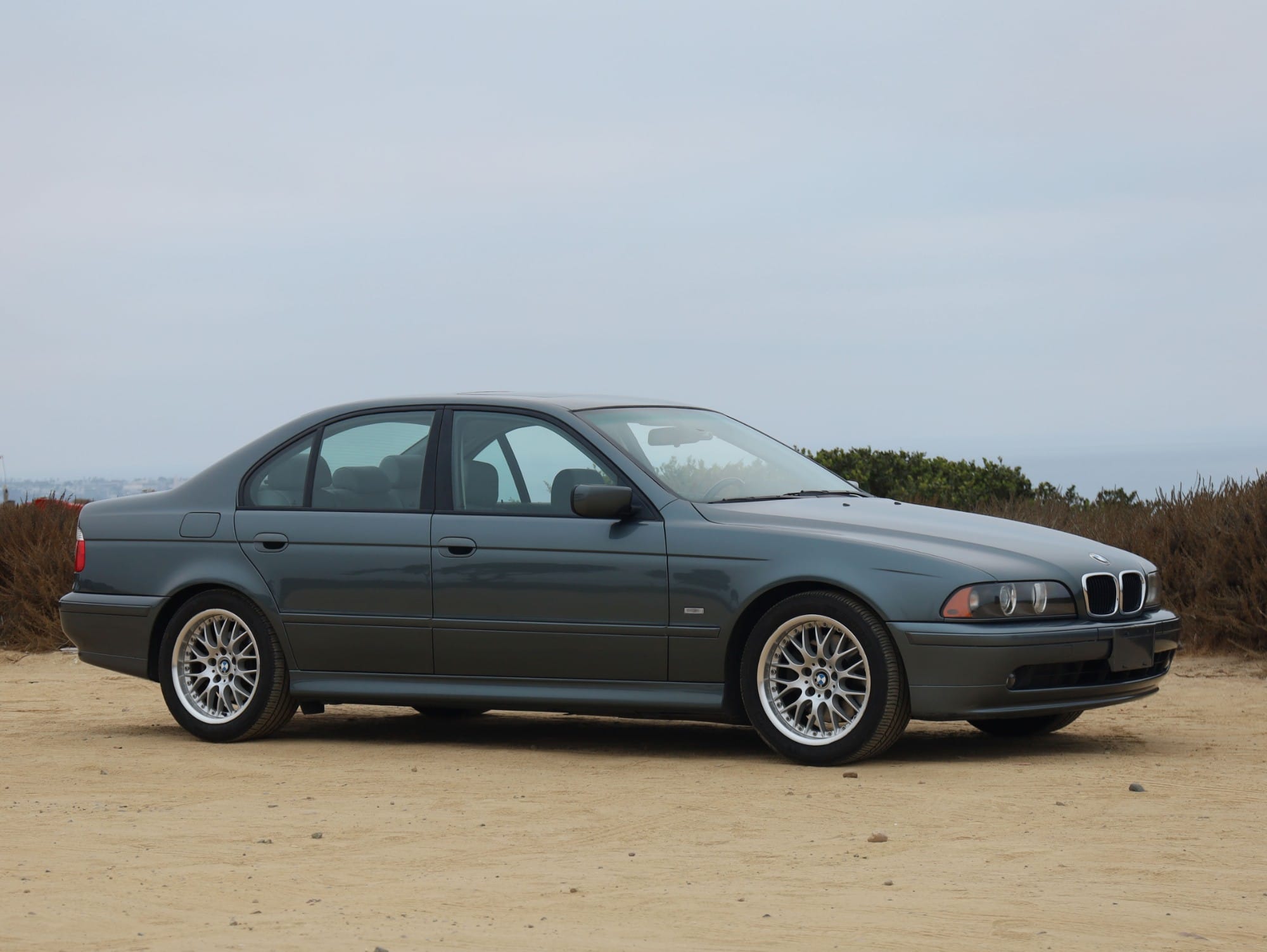 Grey-green 2003 BMW 530i sedan, front three-quarter view on a dirt overlook with coastal scrub behind