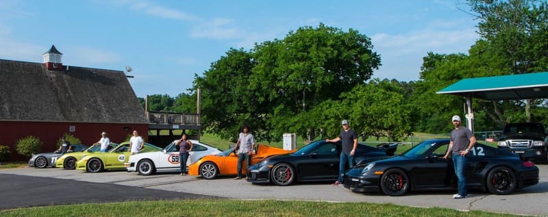 Row of Porsches and other sports cars lined up in a paddock with their owners standing beside them