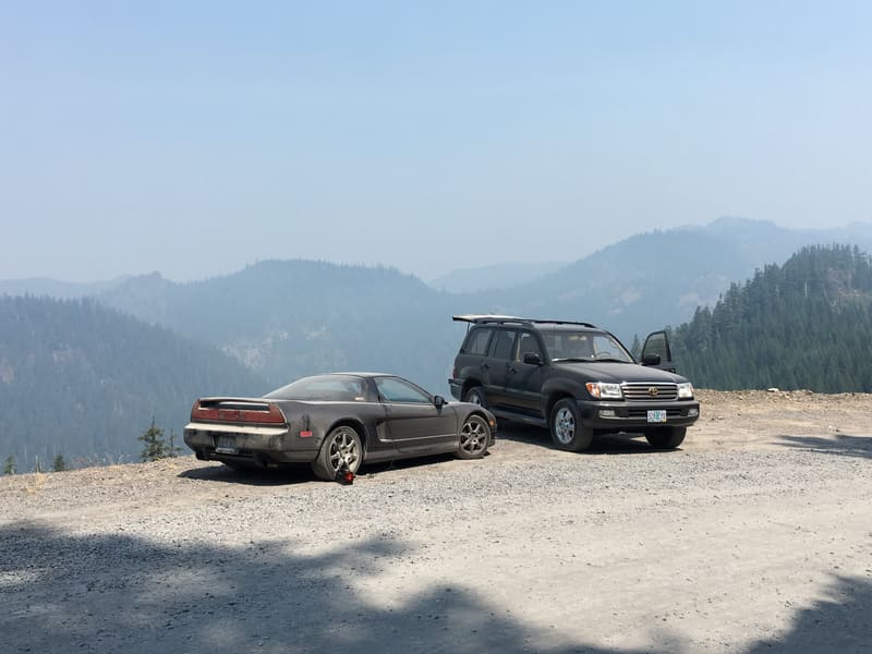 Dark coupe and a Land Cruiser parked at a gravel overlook with smoky mountains behind