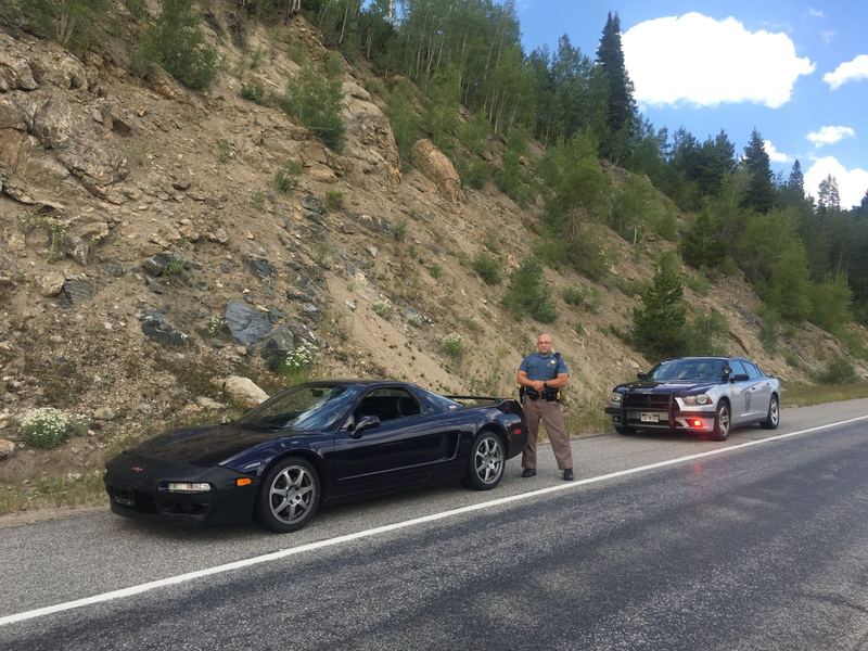 Dark purple Acura NSX pulled over on a mountain road beside a state patrol Dodge Charger, with the trooper standing between the cars