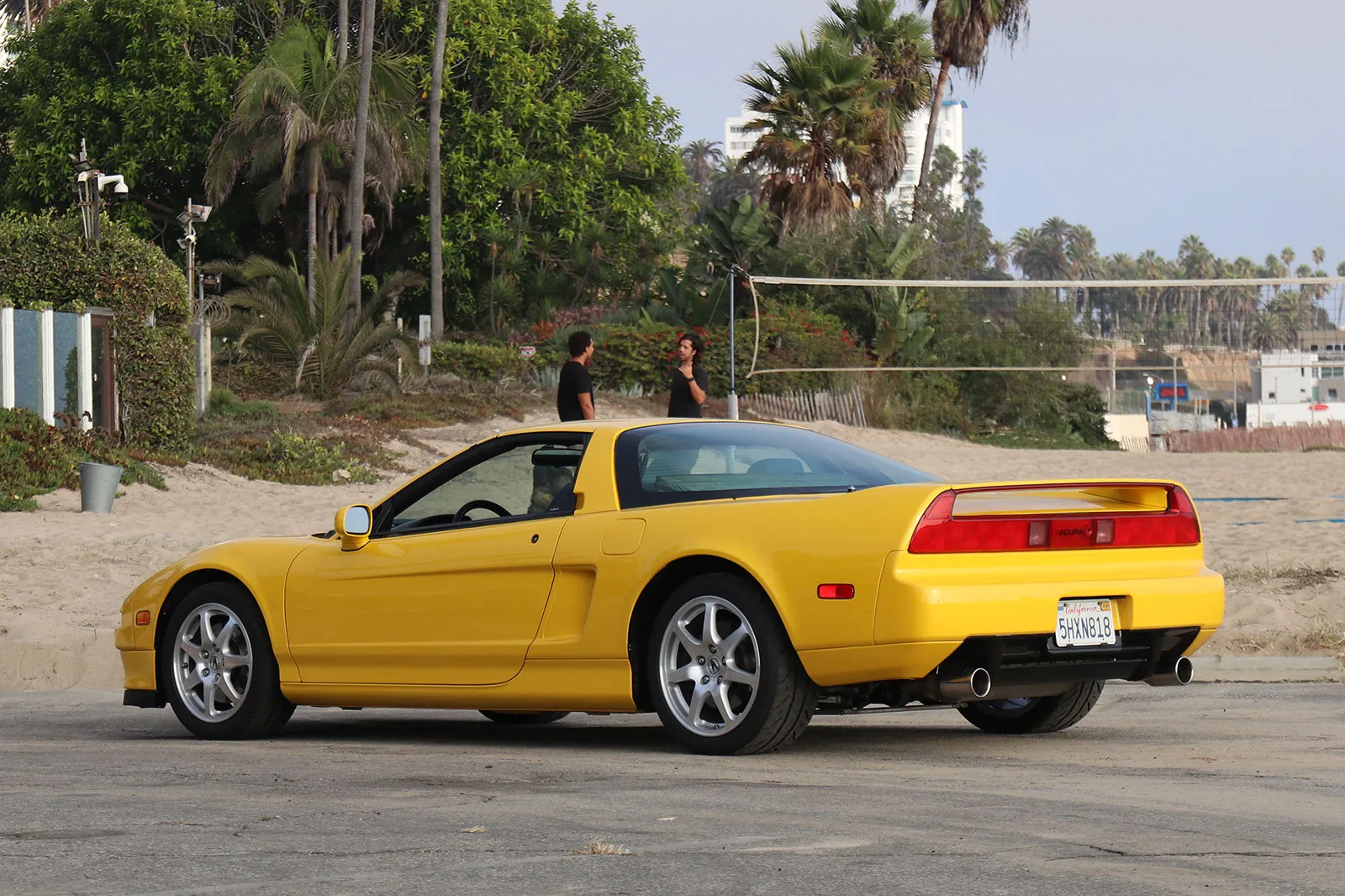 Yellow Acura NSX seen from the rear three-quarter, parked on a beach car park with palm trees behind