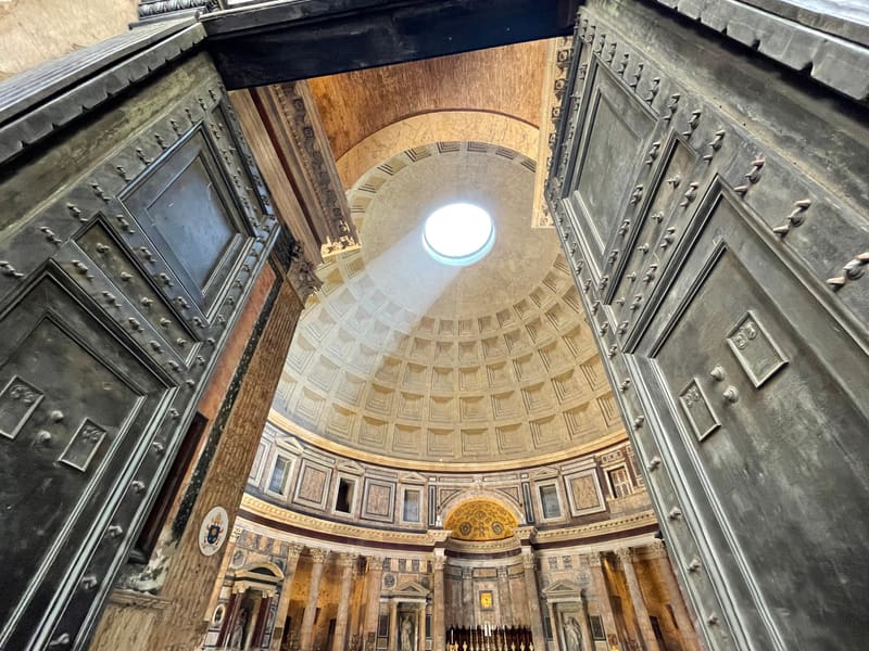 Looking up through large bronze doors into the domed rotunda of the Pantheon in Rome, light coming through the oculus