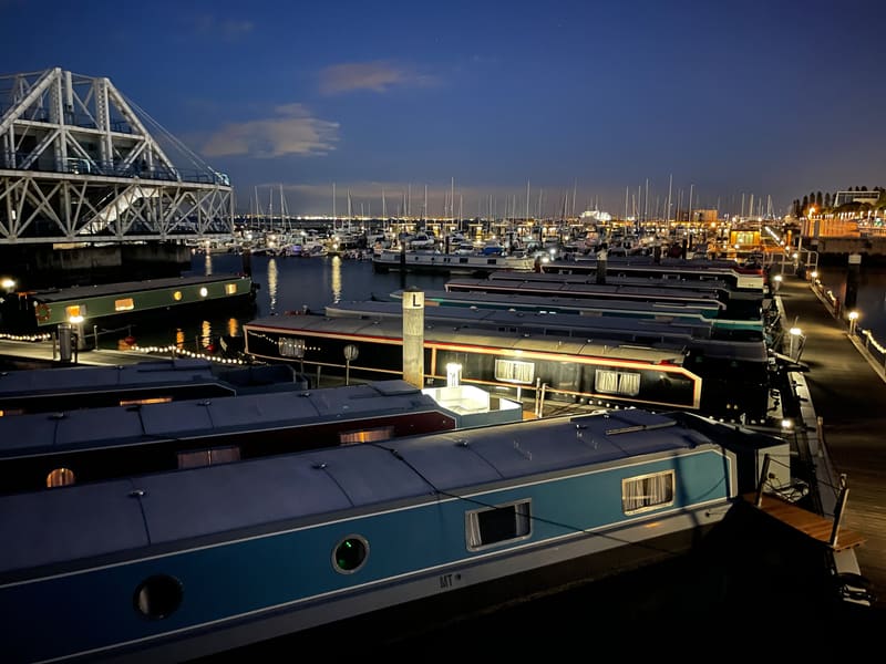 Houseboats moored in a harbor at dusk with a bridge and city lights behind