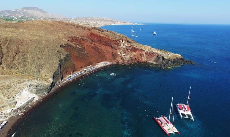 Aerial view of a red-cliffed beach and deep blue water with two catamarans anchored
