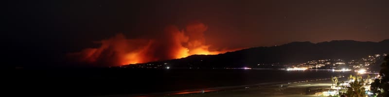 Panorama at night of an orange wildfire glowing behind the Malibu mountains, with the lit Santa Monica Bay shoreline and beach in the foreground